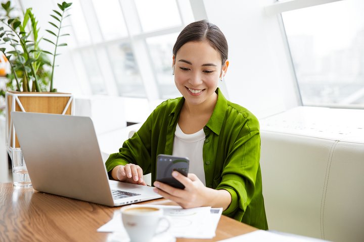 Smiling beautiful young asian woman working on laptop computer building sustainable online presence while sitting at the cafe indoors, using mobile phone